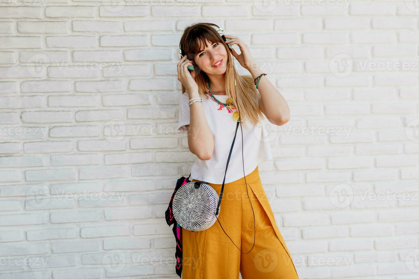 souriant femme posant plus de blanc brique mur avec écouteurs et montrant panneaux. été style. positif ambiance. Jaune des lunettes de soleil. photo