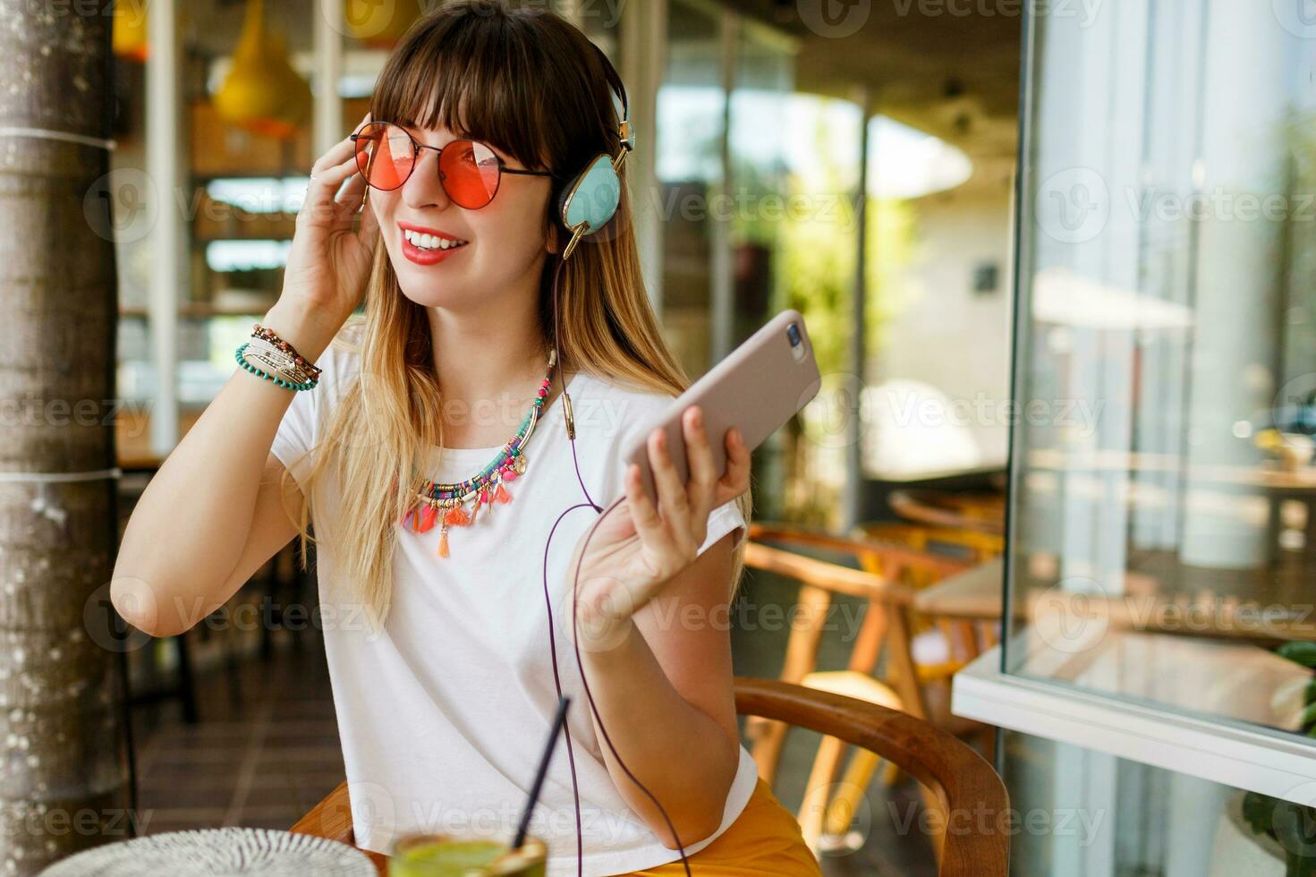 élégant femme dans rose des lunettes profiter vert en bonne santé smoothie , écoute la musique par écouteurs, en portant mobile téléphone. branché accessoires. tropical ambiance. photo
