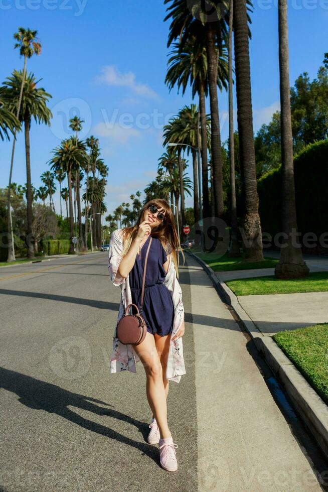 plein longueur portrait de élégant souriant femme en marchant sur exotique rue près le Hôtel dans ensoleillé chaud journée . dépenses sa vacances dans los angeles photo