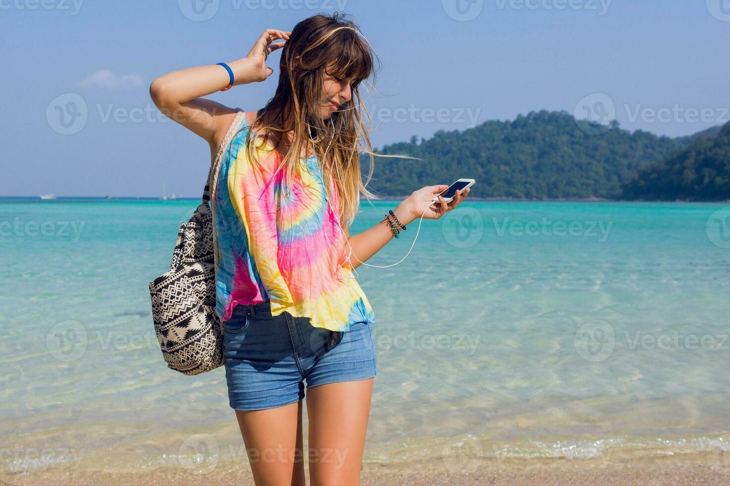 content de bonne humeur Jeune femme écoute à charmant la musique dans écouteurs, en marchant sur le ensoleillé paradis tropical plage. turquoise l'eau Contexte. photo