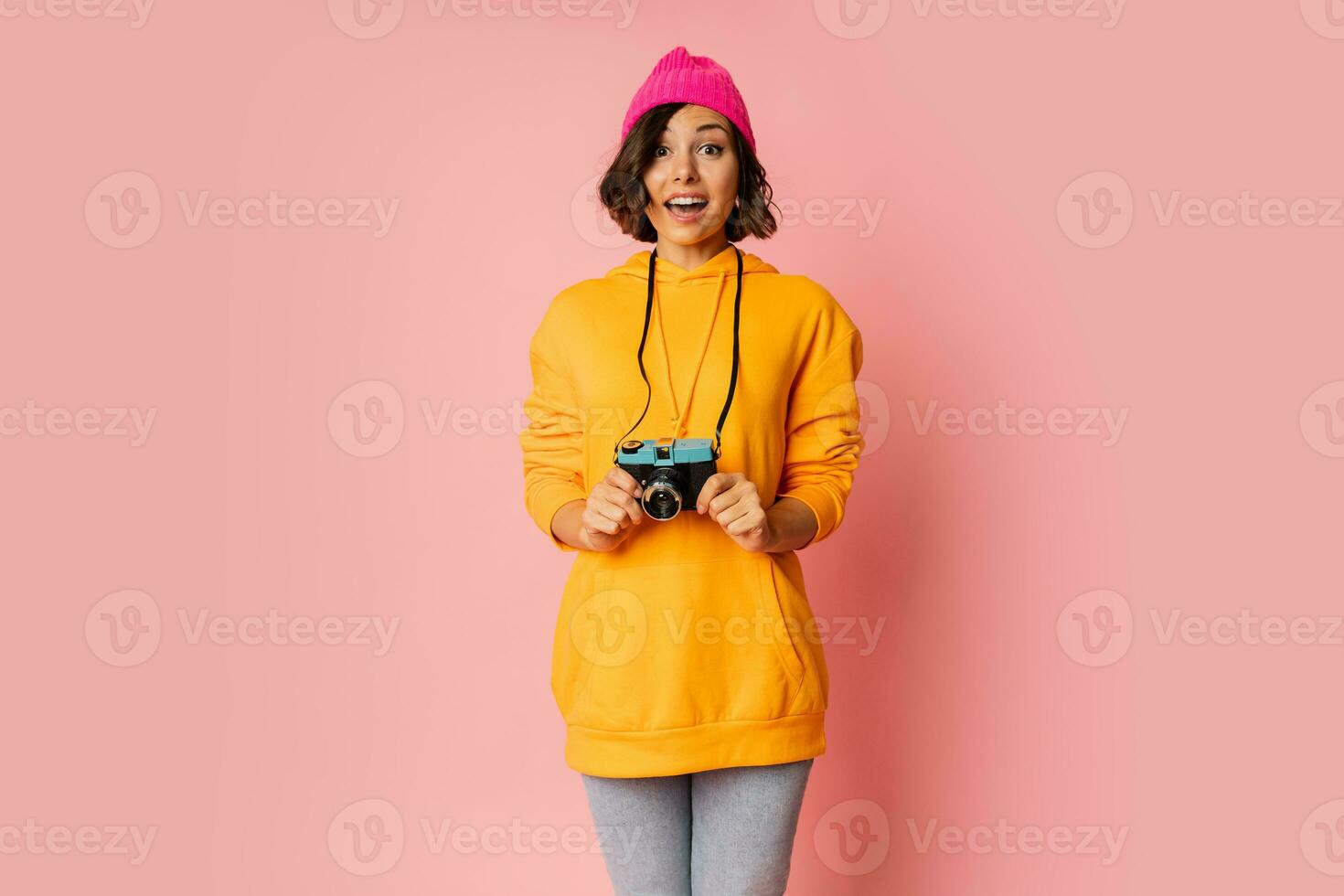 femme dans rose chapeau et Orange sweat à capuche avec surprise visage en utilisant photo caméra sur rose antécédent.