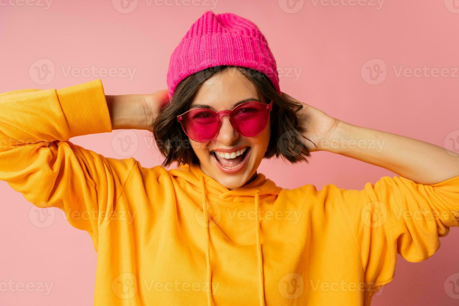 femme dans rose chapeau et Orange sweat à capuche avec émotif visage posant sur rose antécédent. élégant des lunettes de soleil. photo