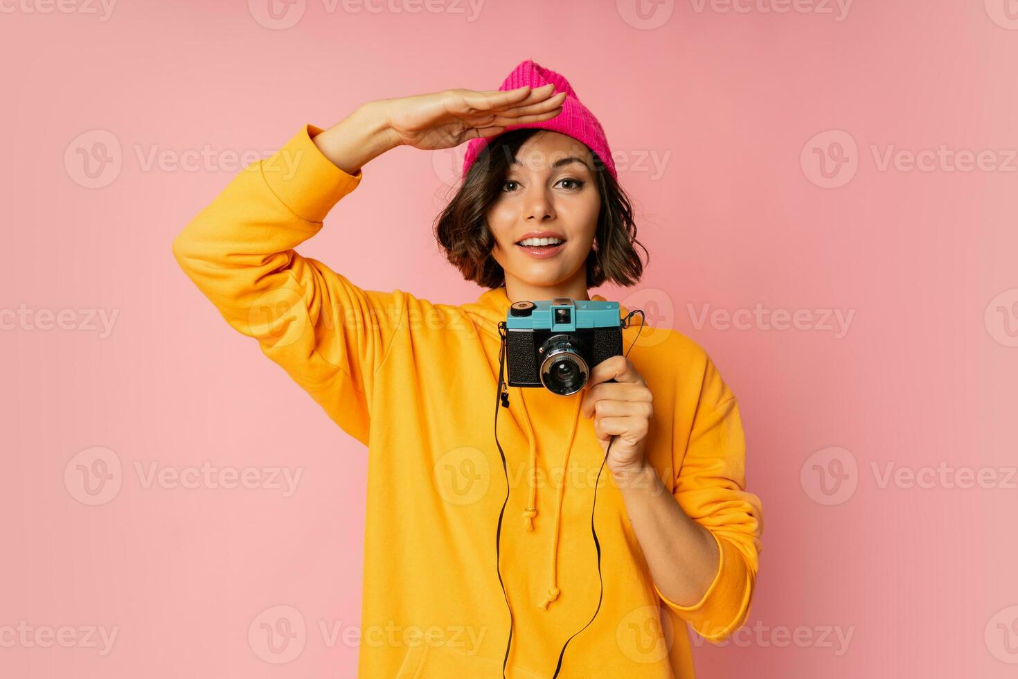 femme dans rose chapeau et Orange sweat à capuche avec surprise visage en utilisant photo caméra sur rose antécédent.