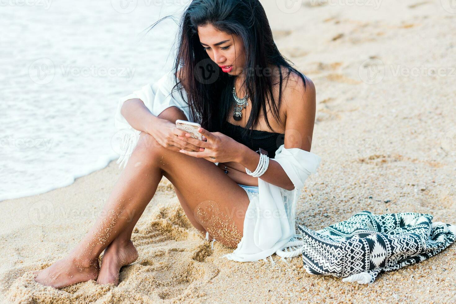 Jeune jolie Asie femme en portant et en utilisant mobile téléphone essai sur tropical plage. portant élégant blanc boho couverture en haut et branché accessoires . rouge lèvres. photo