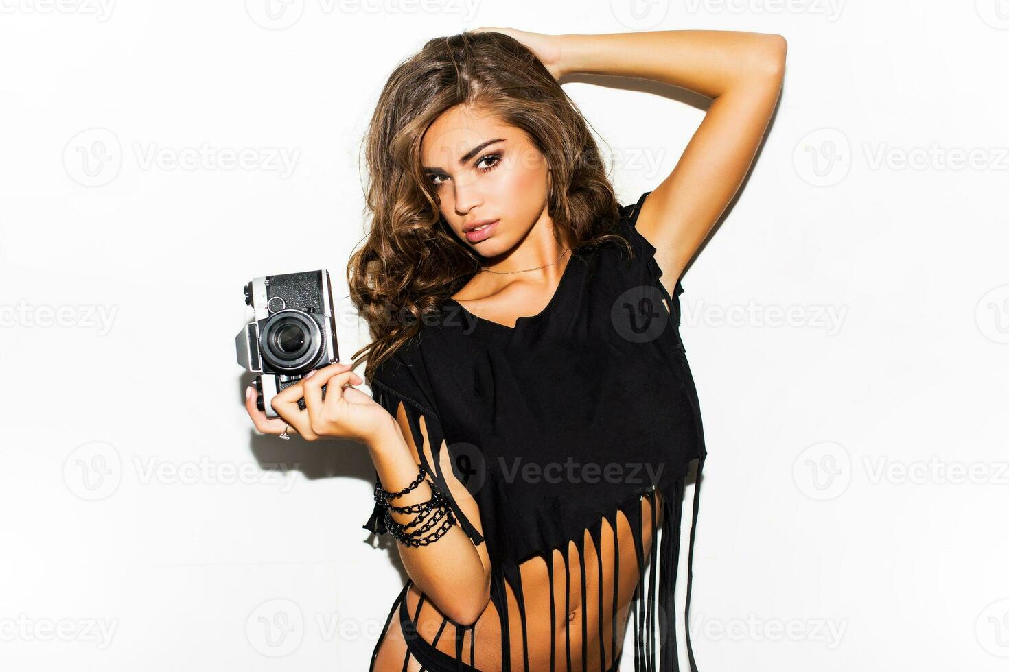 mode de vie image de magnifique sexy Jeune femme dans noir Roche n rouleau T-shirt avec parfait bronzer corps. charme sensuel femme posant avec rétro caméra contre blanc Contexte. intérieur. photo