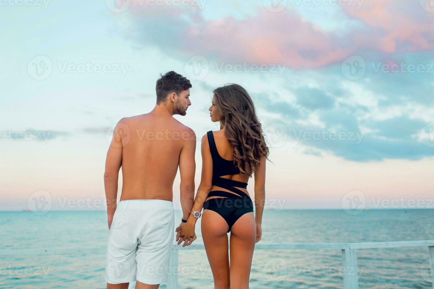 Jeune attrayant couple dans aimer, Beau homme et sexy femme avec parfait bronzer svelte en forme corps profiter romantique soir sur le plage, en portant mains en train de regarder sur le mer. photo