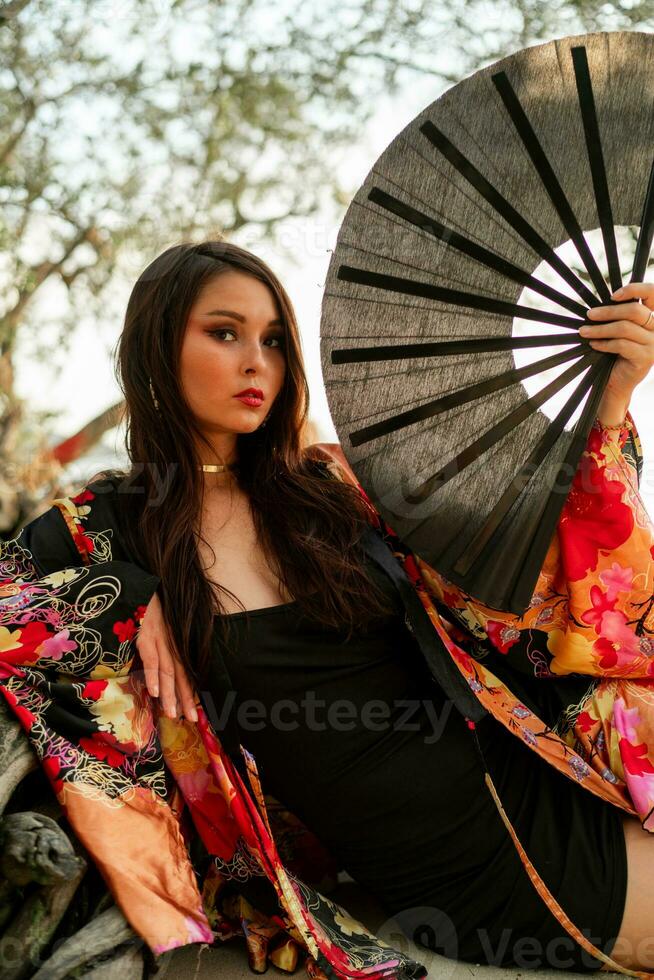 séduisant femme dans élégant kimono avec grand ventilateur et professionnel faire en haut posant Extérieur. photo