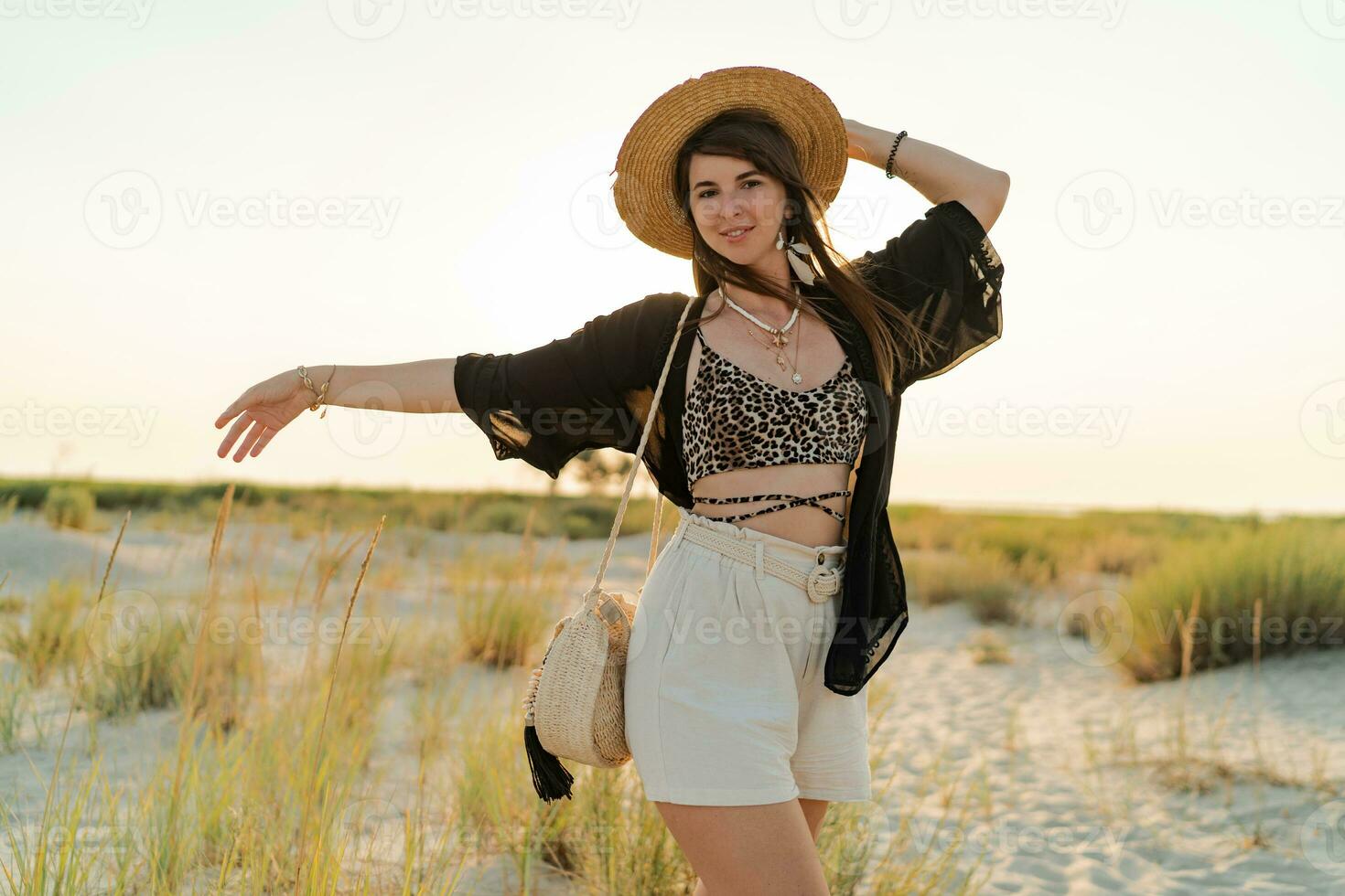 été photo de content Jeune femme dans élégant boho tenue en portant paille sac et chapeau posant sur le l plage. plein longueur.