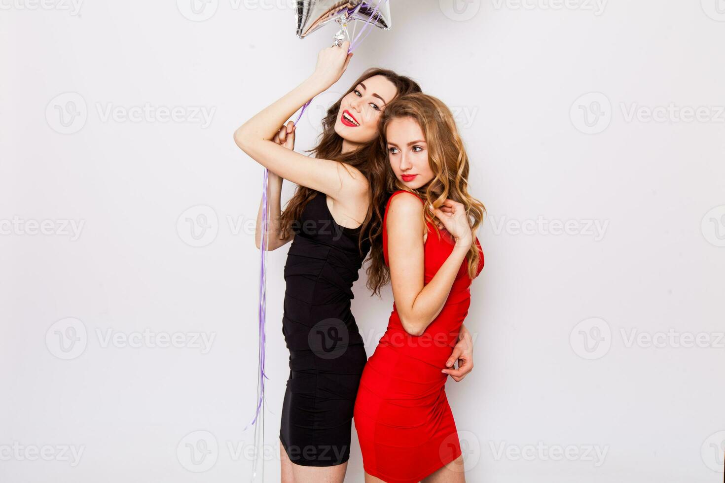 deux magnifique élégant femmes avec rouge lèvres dans soir noir et rouge robe ayant amusement. un en gardant rouge étoiles des ballons dans sa main et souriant. deux femme à le faire la fête. à l'intérieur. blanc Contexte. photo