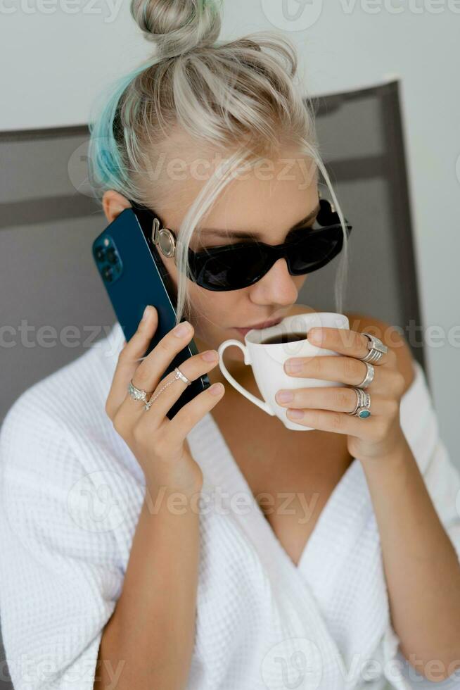 proche en haut portrait de souriant blond femme dans élégant des lunettes de soleil et blanc peignoir en buvant tasse de café dans de bonne heure Matin. repos dans luxe Hôtel. photo