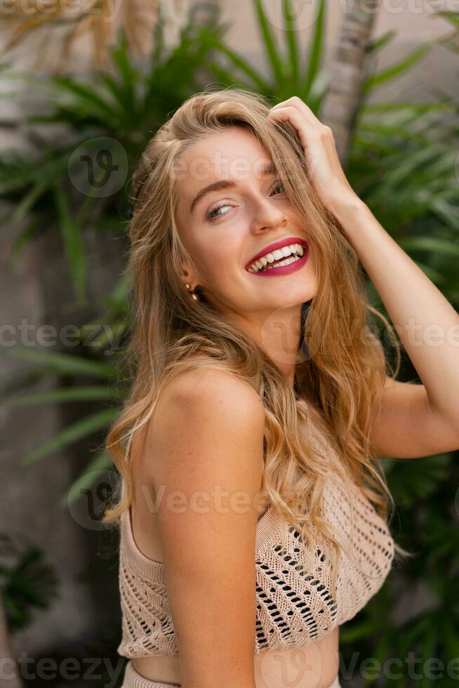 content en riant blond femme avec ondulé Cheveux posant dans élégant tropical villa. vacances ambiance. rouge lèvres, parfait sourire. photo