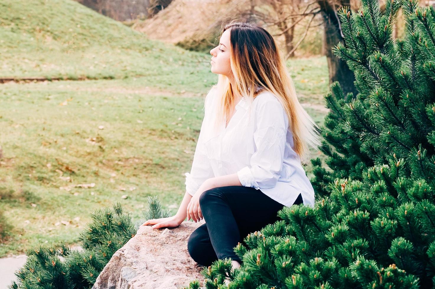 un portrait d'une jolie jeune fille aux cheveux longs en chemise blanche, assise sur une pierre dans un parc, avec fond d'herbe verte et pins au premier plan photo