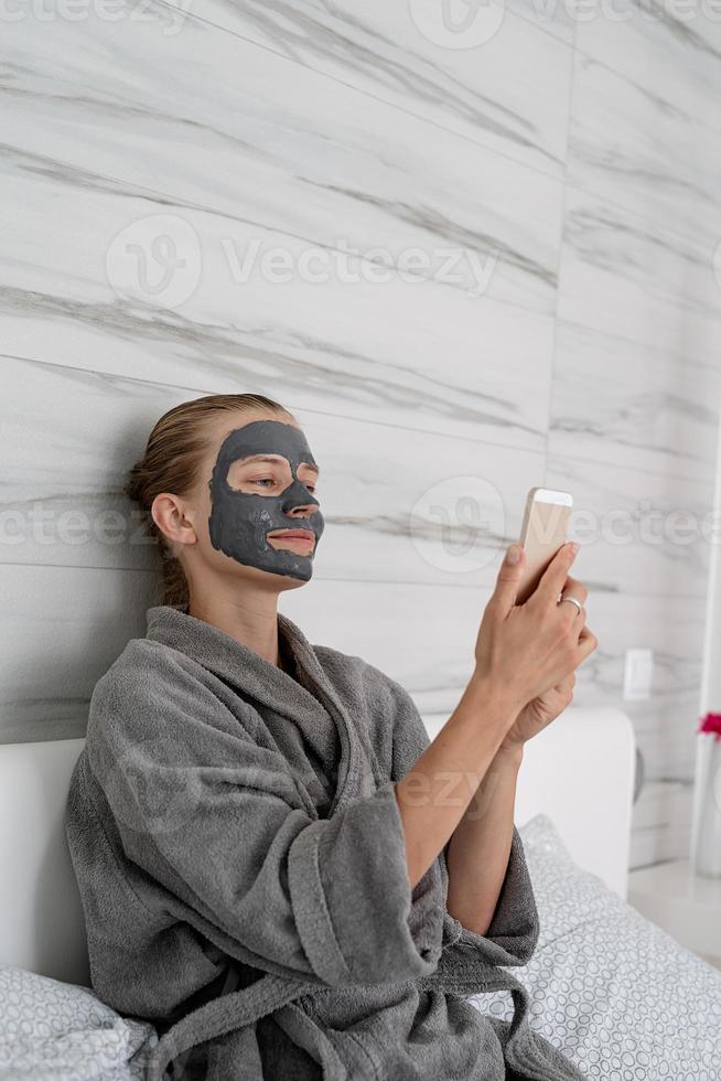 femme avec masque facial relaxant assise sur le lit à l'aide d'un appareil mobile photo