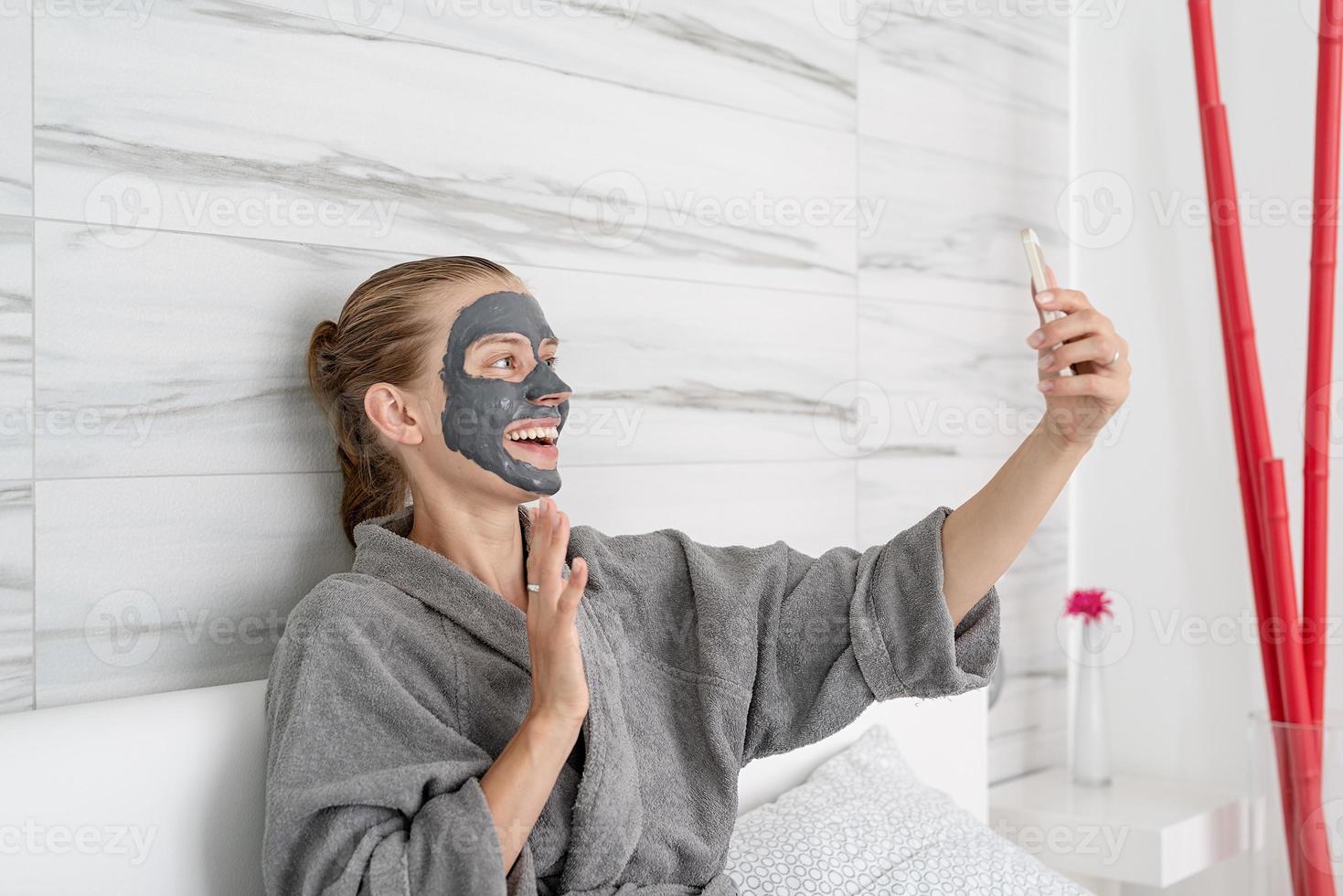 femme avec masque facial relaxant assise sur le lit à l'aide d'un appareil mobile photo
