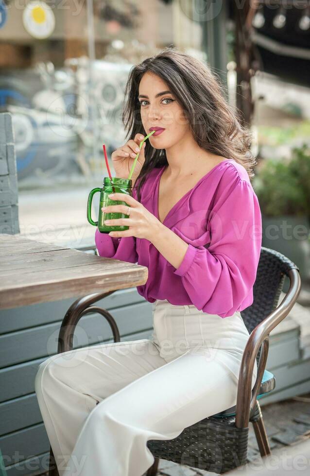 content brunette fille avec rose chemisier séance à le bar , en buvant une verre de limonade tandis que souriant à le caméra. Jeune sexy jolie femme sur le chaise en buvant jus portant une blanc un pantalon photo