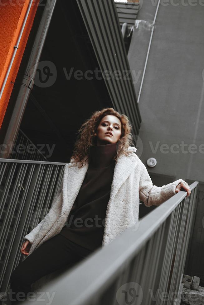 une fille aux cheveux bouclés rouges dans un manteau blanc pose sur les escaliers du parking photo