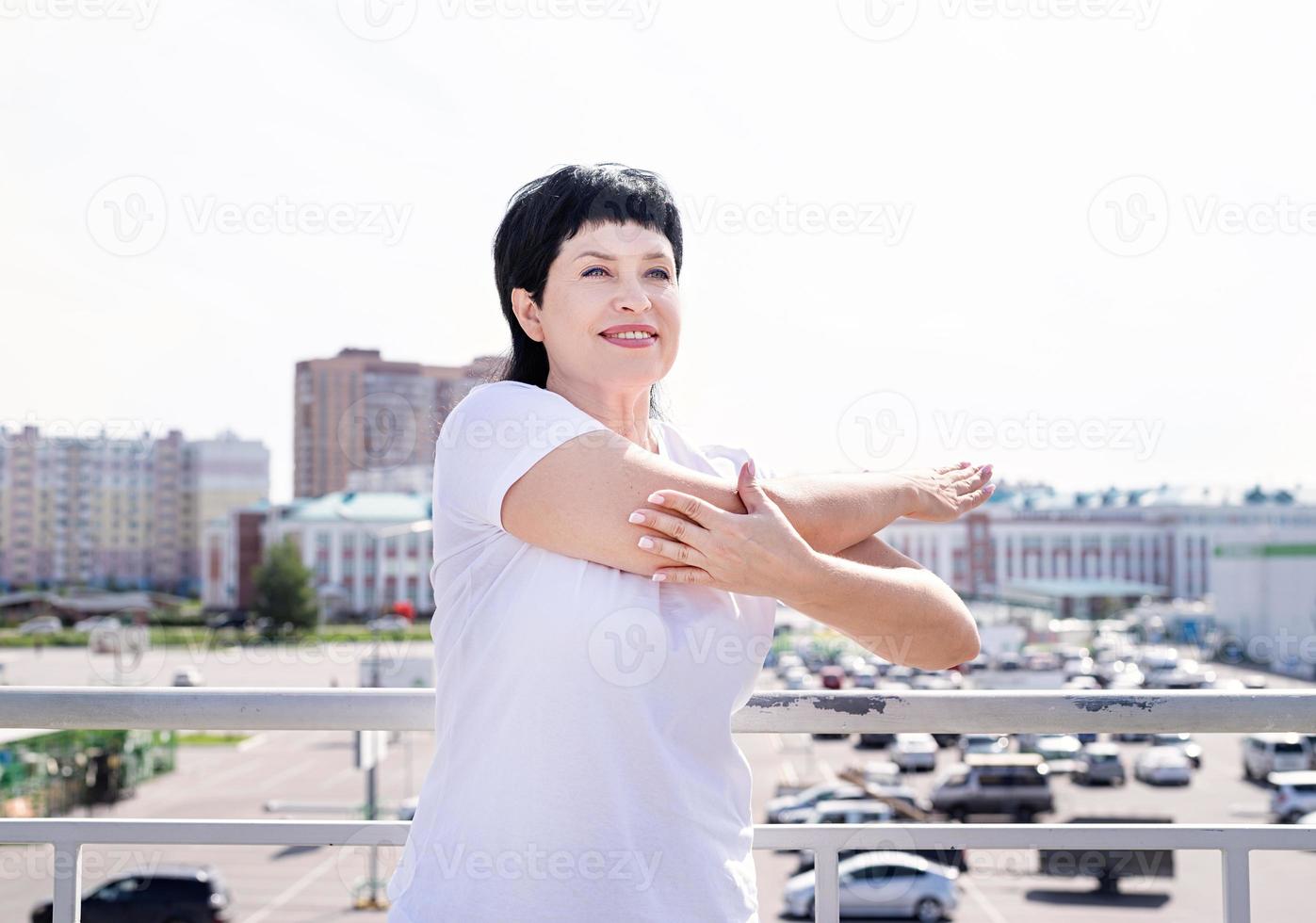 femme âgée souriante faisant des étirements à l'extérieur sur fond urbain photo