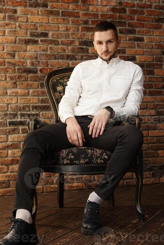 bel homme à la mode dans une chemise blanche est assis sur une chaise dans un studio photo