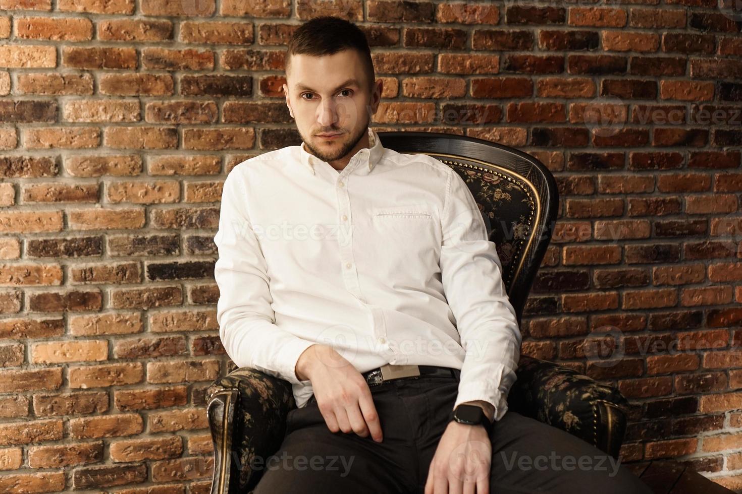 bel homme à la mode dans une chemise blanche est assis sur une chaise dans un studio photo