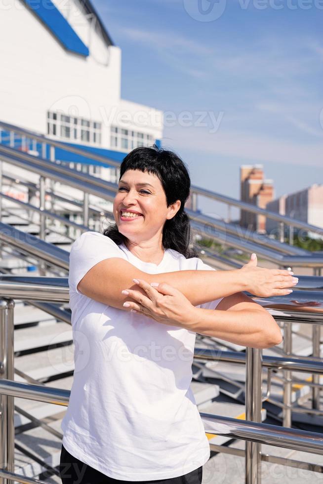 femme âgée souriante faisant des étirements à l'extérieur sur fond urbain photo