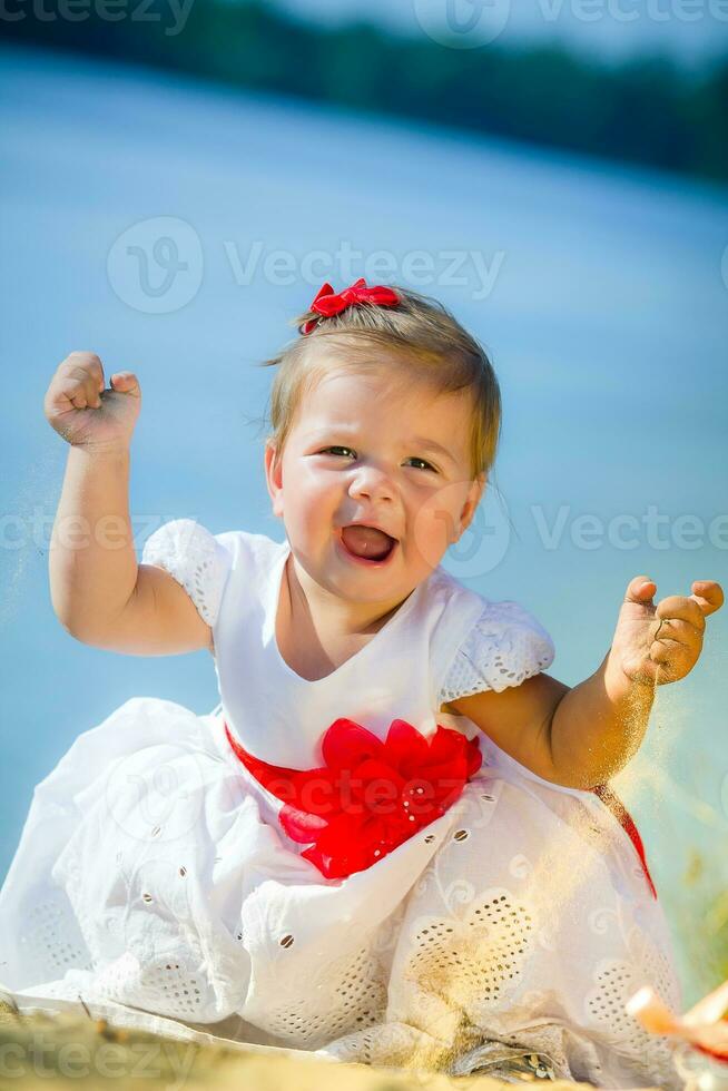 le peu fille dans une blanc robe avec une gros rouge arc est assis sur le bord de la rivière. bébé pleure, élevage le sien mains en haut. enfant verse le sable de le sien mains sur le plage. photo