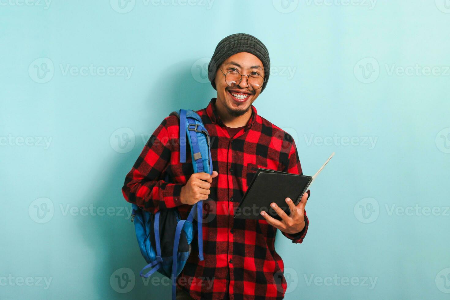 Jeune asiatique étudiant homme portant une sac à dos est en portant livres tandis que permanent contre une bleu Contexte photo