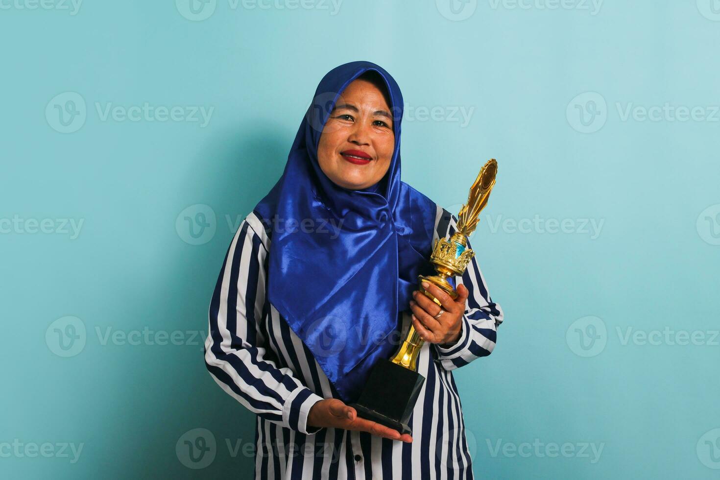 un excité âge moyen asiatique femme d'affaires dans une bleu hijab et une rayé chemise est en portant une or trophée, célébrer sa Succès et réalisation. elle est isolé sur une bleu Contexte photo