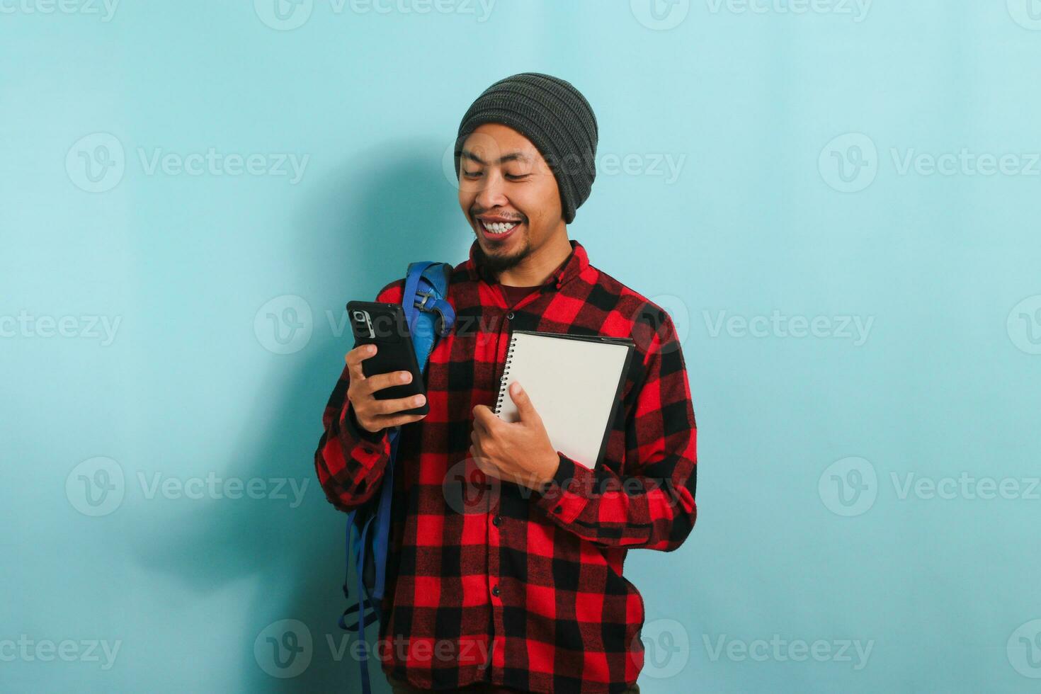 un excité Jeune asiatique Masculin étudiant, portant une sac à dos, bonnet chapeau, et rouge plaid flanelle chemise, détient une livre dans un main et les usages le sien téléphone avec le autre, tandis que permanent contre une bleu Contexte photo