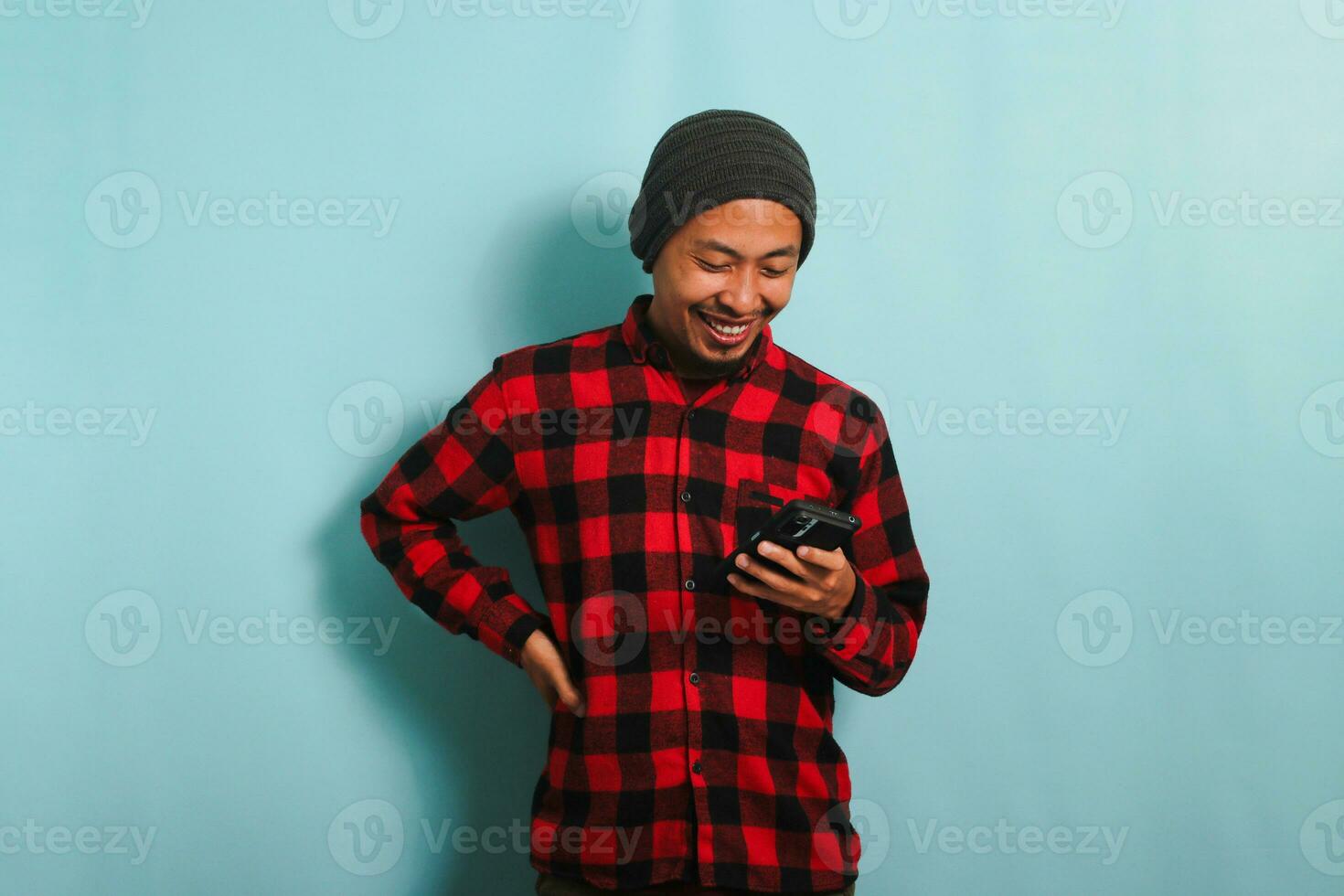 content Jeune asiatique homme avec une bonnet chapeau et une rouge plaid flanelle chemise est en utilisant une téléphone intelligent à vérifier social médias et magasin en ligne, isolé sur une bleu Contexte photo