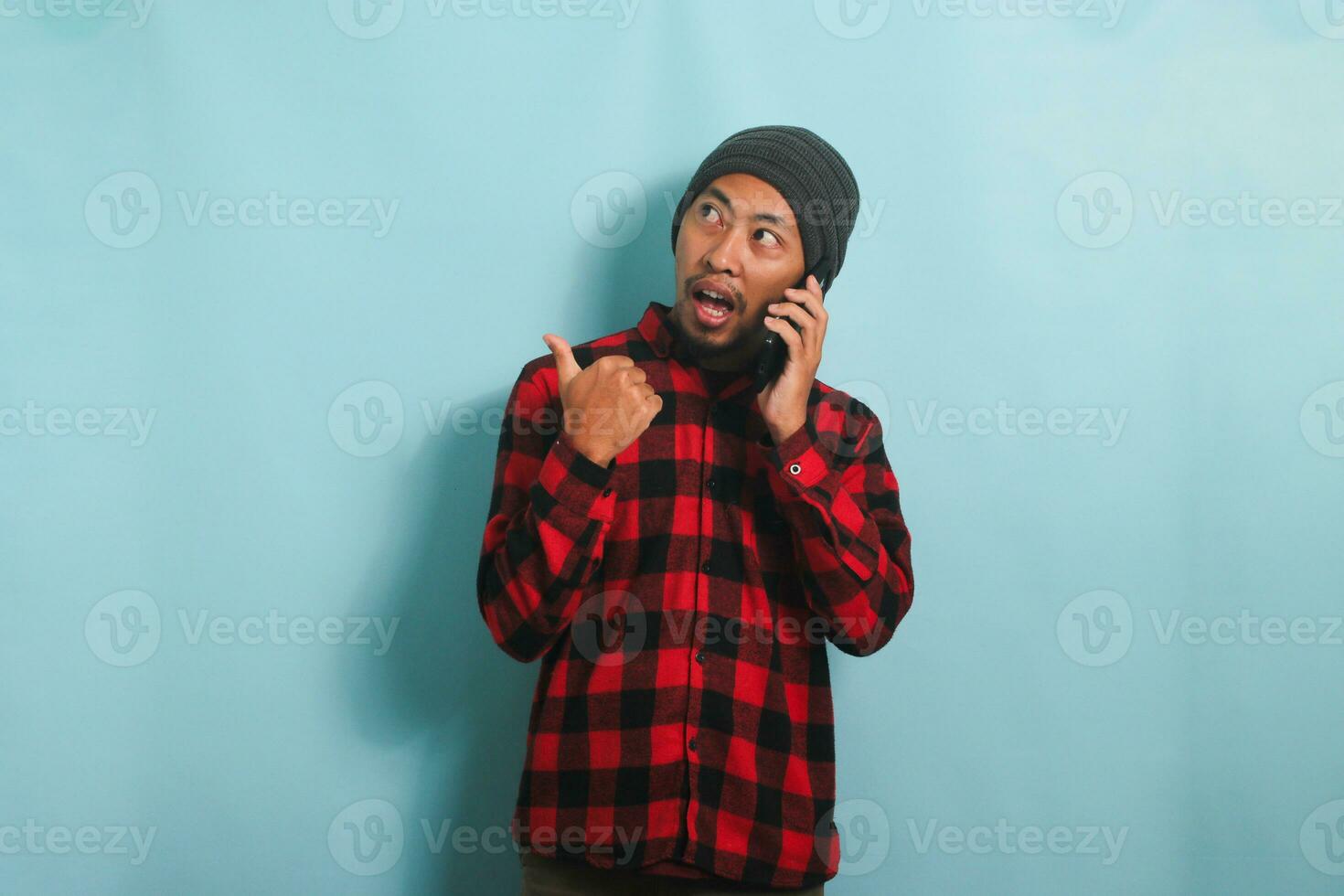 excité Jeune asiatique homme avec une bonnet chapeau et une rouge plaid flanelle chemise est montrer du doigt le sien doigt à le la gauche à copie espace avec pouce tandis que parlant sur le sien mobile téléphone, isolé sur une bleu Contexte photo