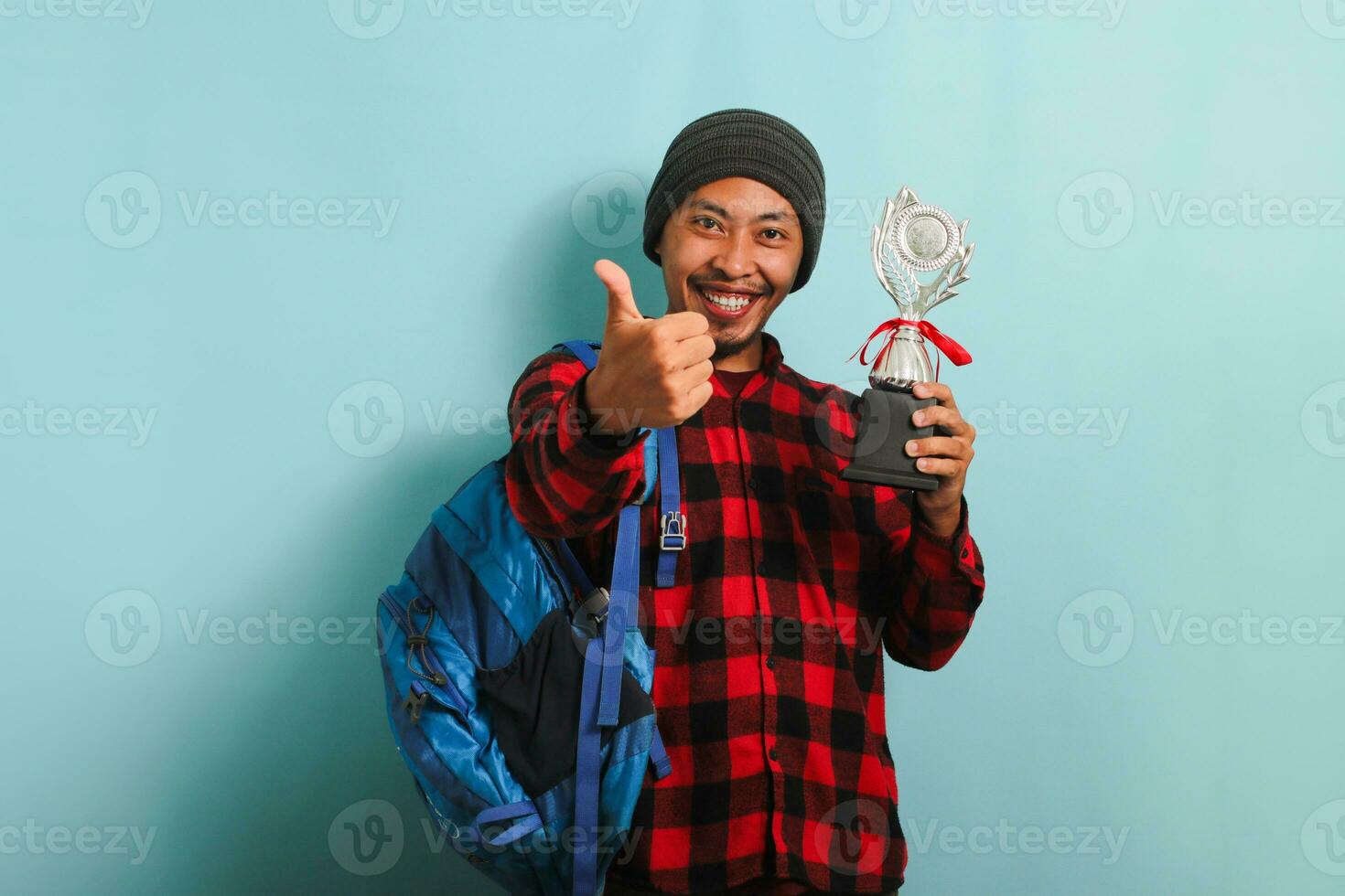 excité Jeune asiatique homme étudiant portant sac à dos, bonnet chapeau, et rouge plaid flanelle chemise, donnant pouces vers le haut tandis que en portant trophée, réjouissance dans le sien Succès et réalisation, isolé sur bleu Contexte photo