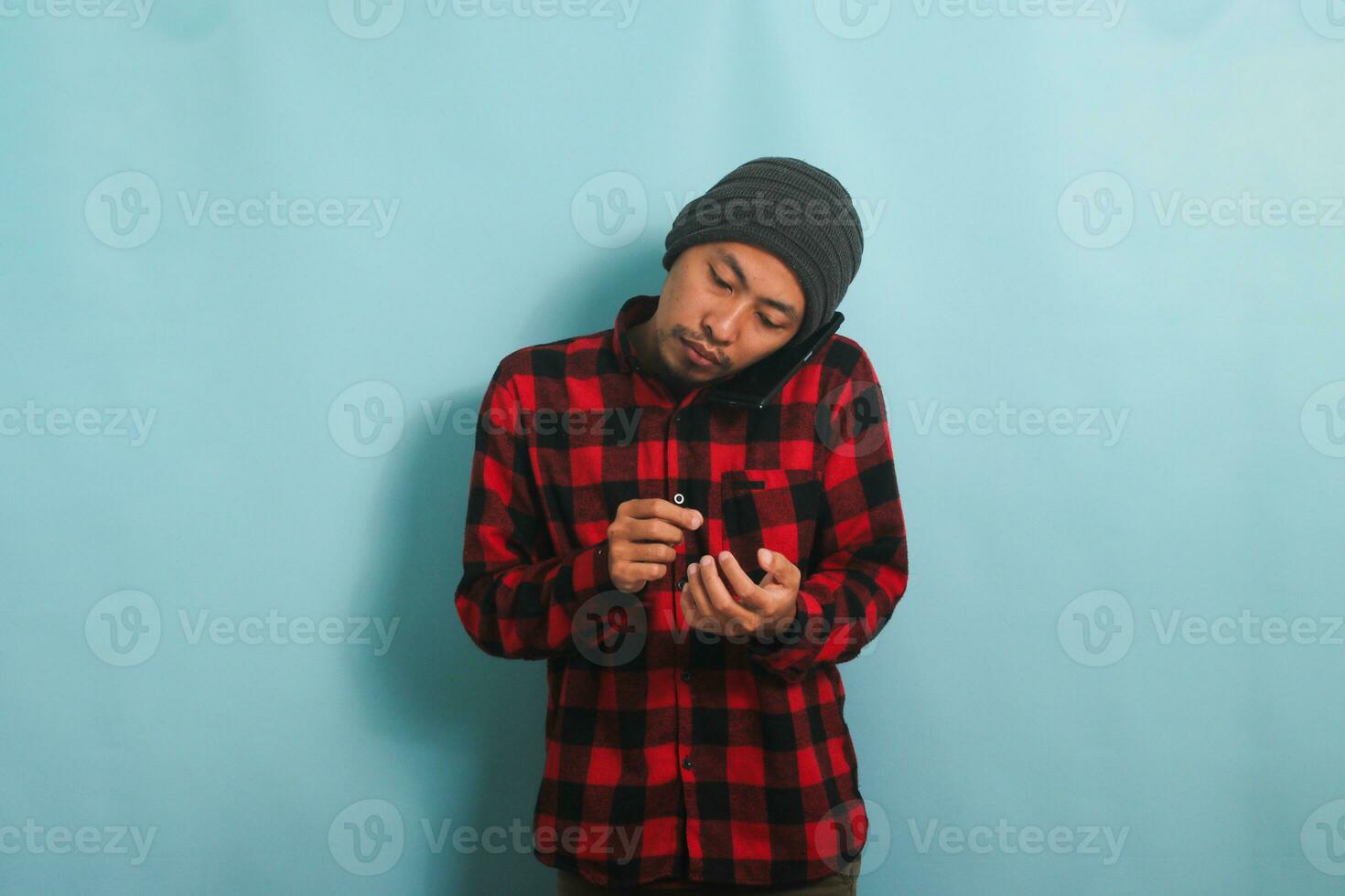 Jeune asiatique homme avec une bonnet chapeau et une rouge plaid flanelle chemise est fabrication Remarques tandis que parlant sur le sien mobile téléphone, isolé sur une bleu Contexte photo