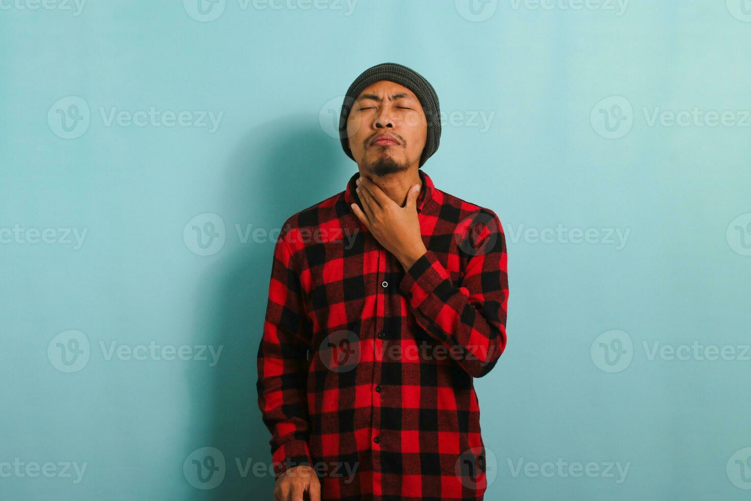 Jeune asiatique homme avec bonnet chapeau et rouge plaid flanelle chemise émouvant le sien douloureux cou, Souffrance de une endolori gorge pendant grippe saison, isolé sur une bleu Contexte photo