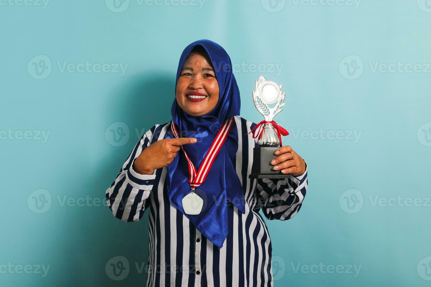 une content âge moyen asiatique femme d'affaires dans une bleu hijab, rayé chemise, et médaille est montrer du doigt une argent trophée, célébrer sa Succès et réalisation, isolé sur une bleu Contexte photo