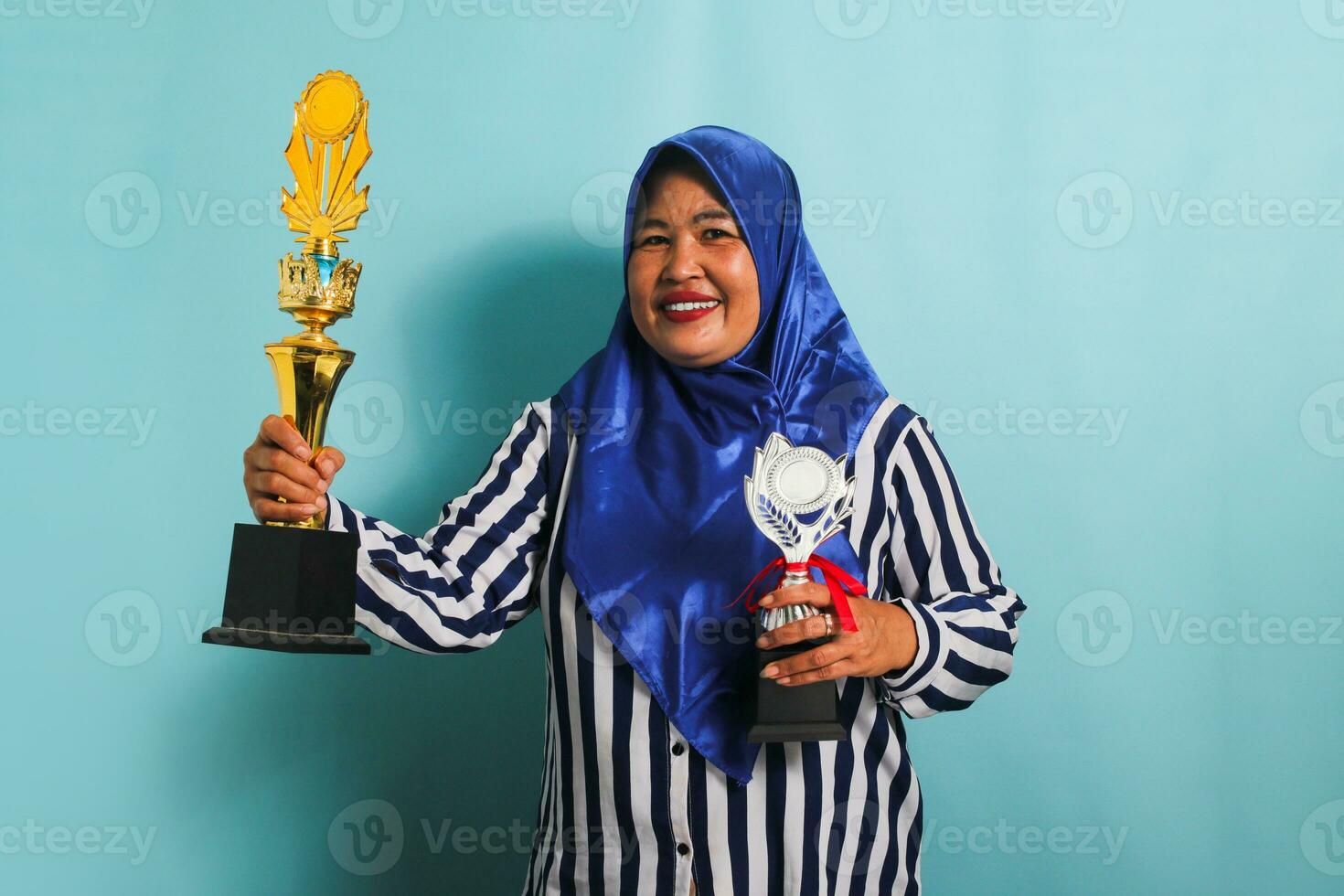 un excité âge moyen asiatique femme d'affaires dans une bleu hijab et une rayé chemise est en portant une or et argent trophée, célébrer sa Succès et réalisation. elle est isolé sur une bleu Contexte photo