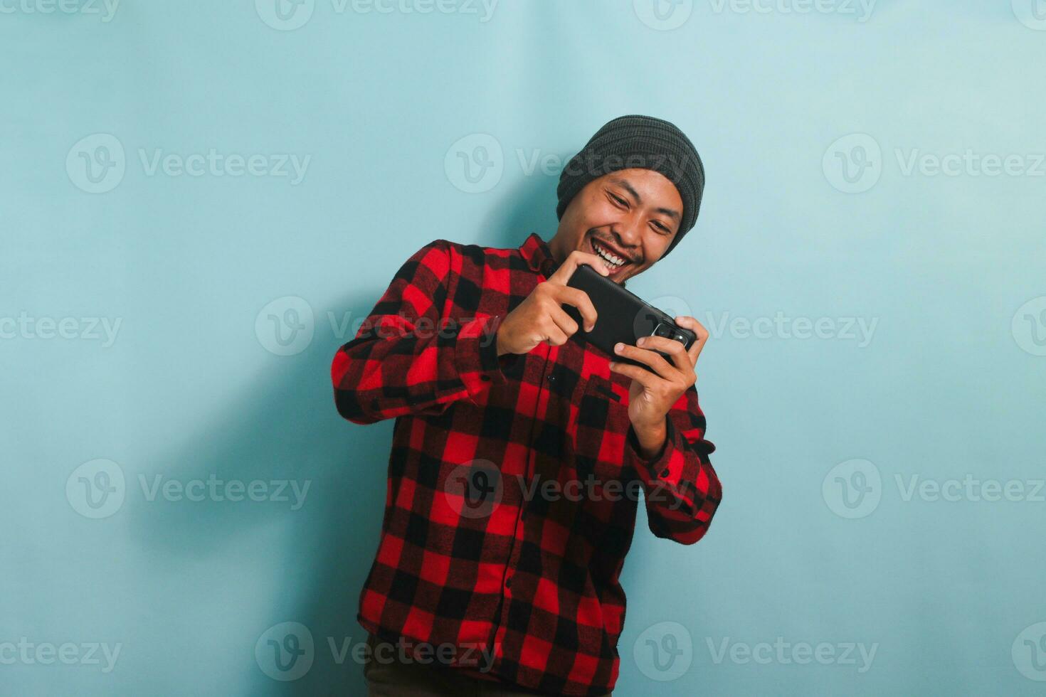 excité Jeune asiatique homme avec une bonnet chapeau et une rouge plaid flanelle chemise est en jouant une courses Jeu sur le sien téléphone intelligent, isolé sur une bleu Contexte photo