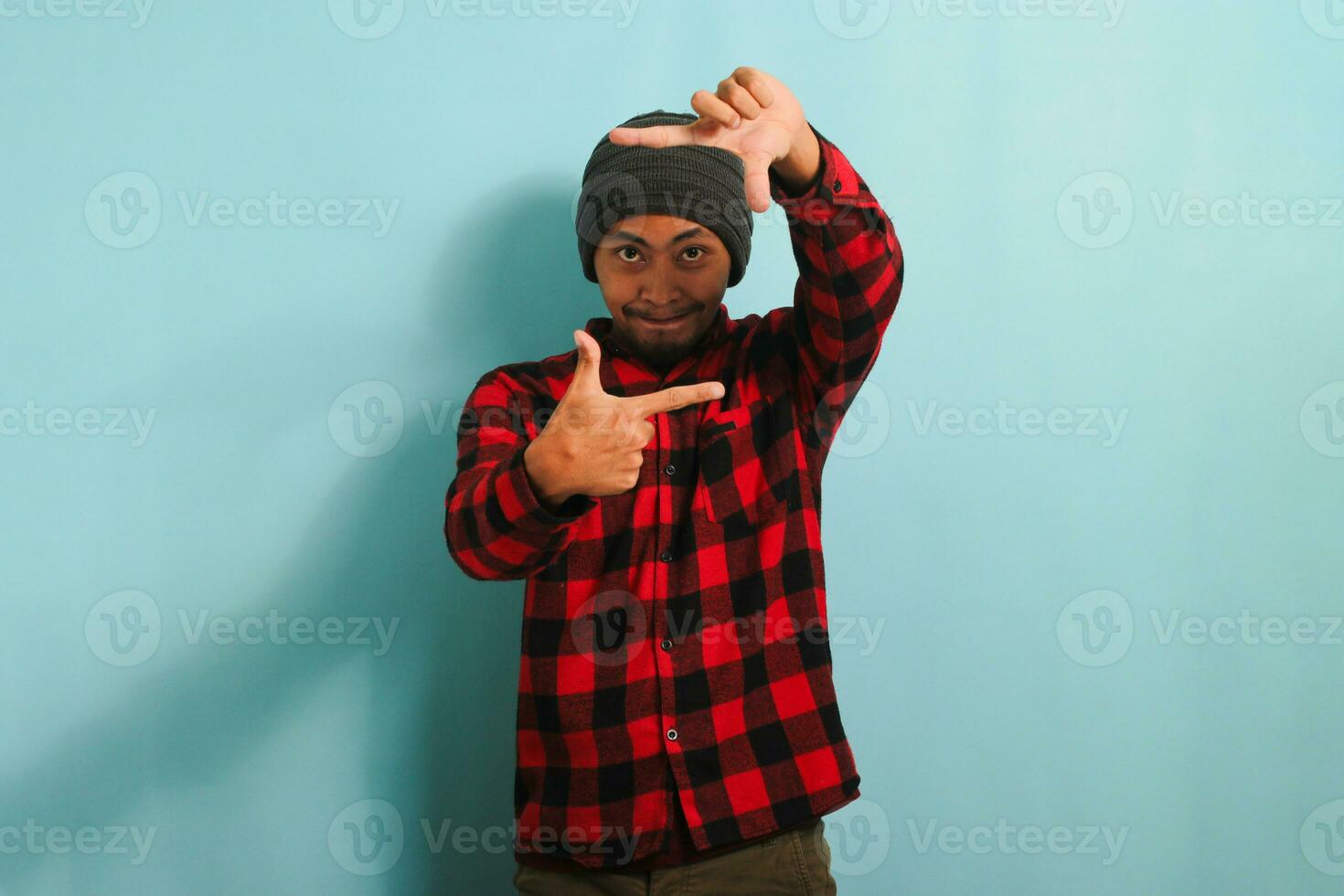 excité Jeune asiatique homme avec une bonnet chapeau et une rouge plaid flanelle chemise est fabrication une image Cadre geste, prétendre à concentrer comme une photographe, isolé sur une bleu Contexte photo