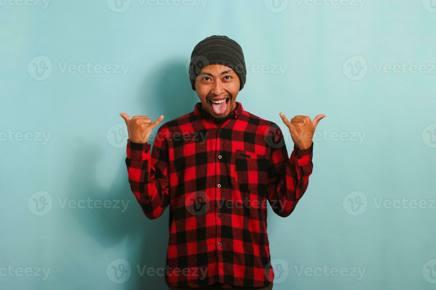 content Jeune asiatique homme avec une bonnet chapeau et rouge plaid flanelle chemise montrant le Roche et rouleau geste avec le sien les doigts et collage en dehors le sien langue, isolé sur une bleu Contexte photo