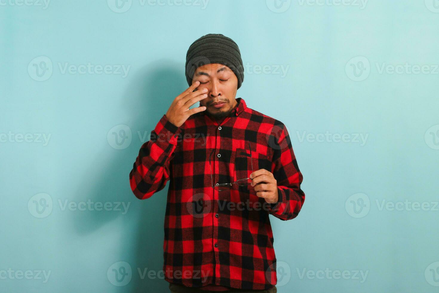 Jeune asiatique homme avec bonnet chapeau et rouge plaid flanelle chemise expérience malaise avec contact lentilles, frottement le sien gonflé yeux dû à poussière allergie et démangeaison, isolé sur une bleu Contexte photo