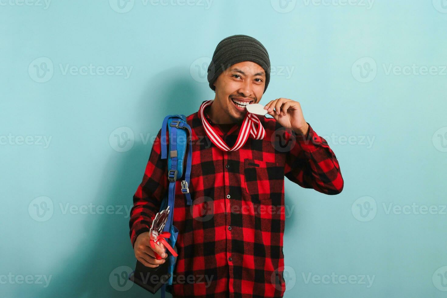 content Jeune asiatique homme étudiant portant une sac à dos, bonnet chapeau, et rouge plaid flanelle chemise, mordant une médaille tandis que en portant une trophée, réjouissance dans Succès et réalisation, isolé sur une bleu Contexte photo