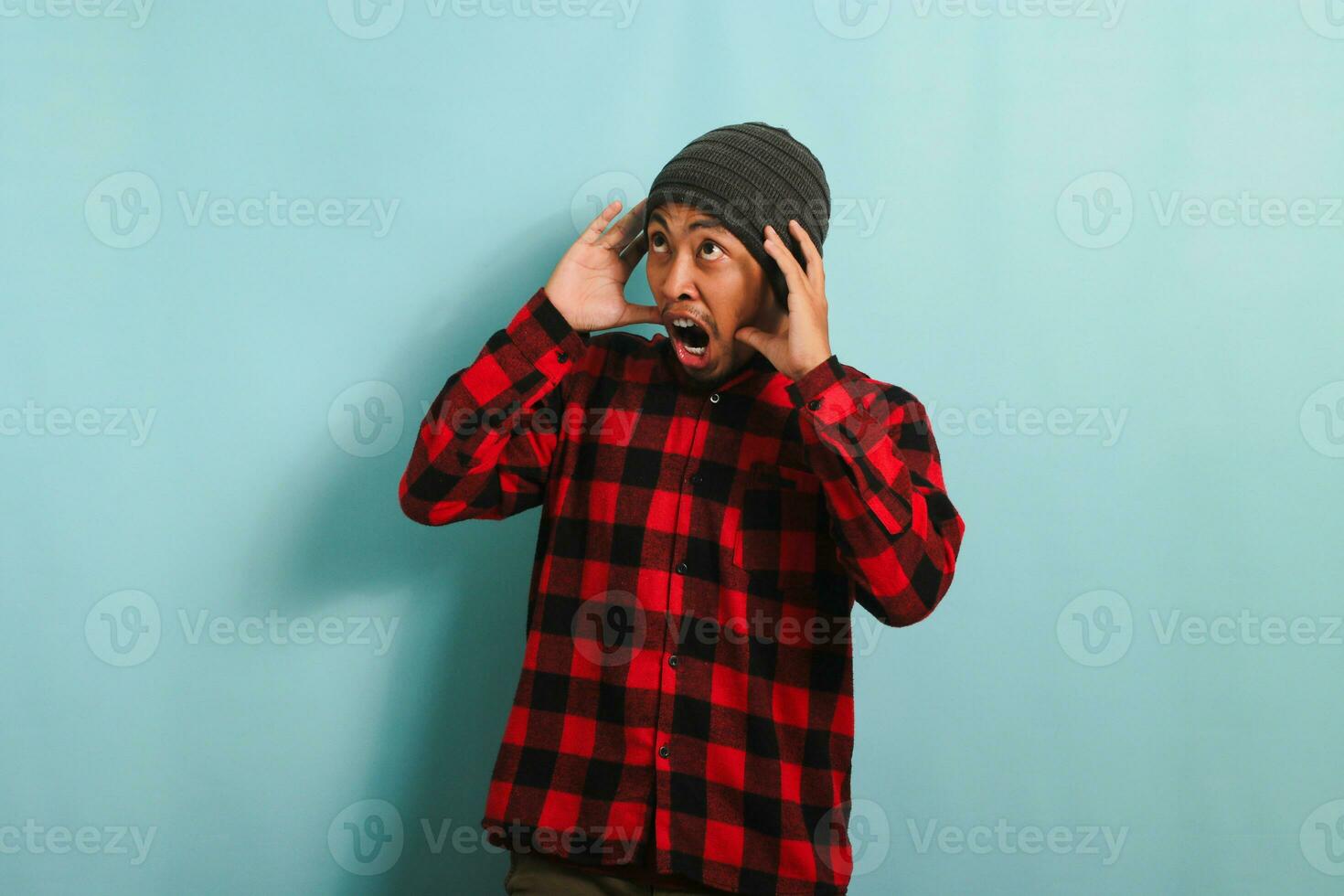 sous le choc Jeune asiatique homme avec une bonnet chapeau et rouge plaid flanelle chemise touche le sien diriger, à la recherche de côté à copie espace dans incrédulité, isolé sur une bleu Contexte photo