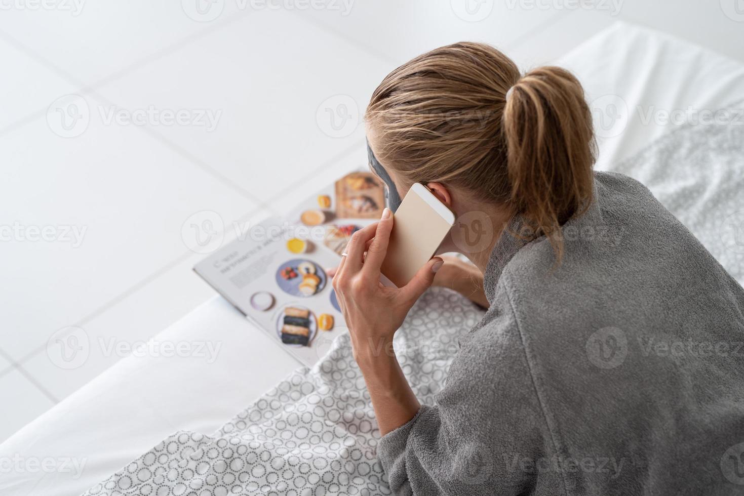 femme avec masque facial relaxant allongé sur le lit en lisant un magazine photo