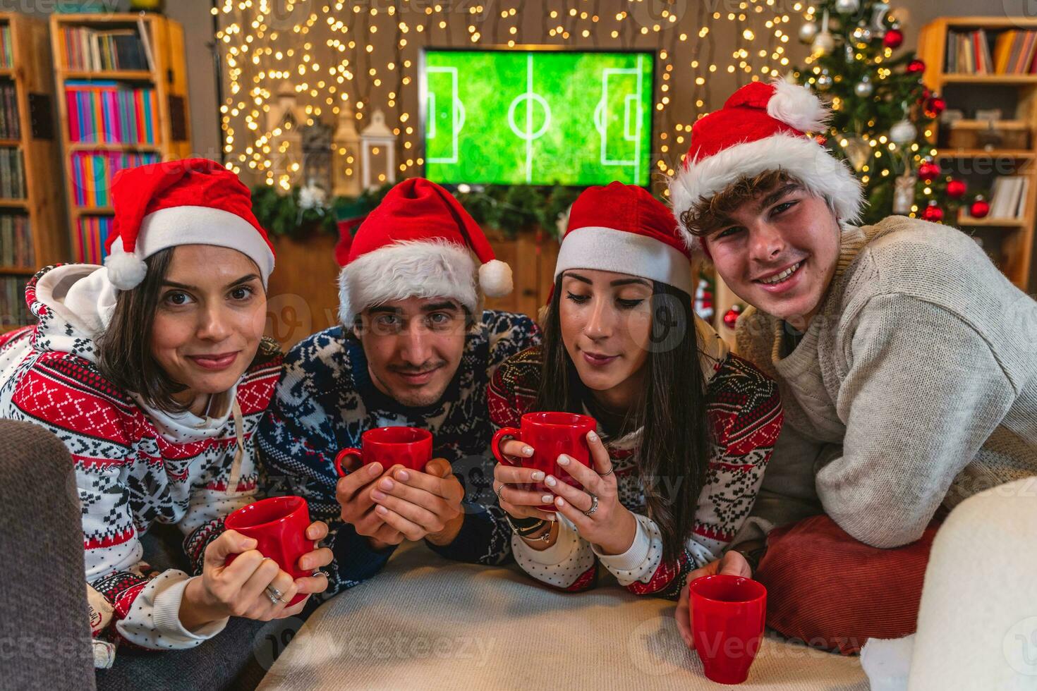 groupe de quatre Jeune copains grillage avec rouge tasses habillé dans rouge Chapeaux et Noël pulls molletonnés photo