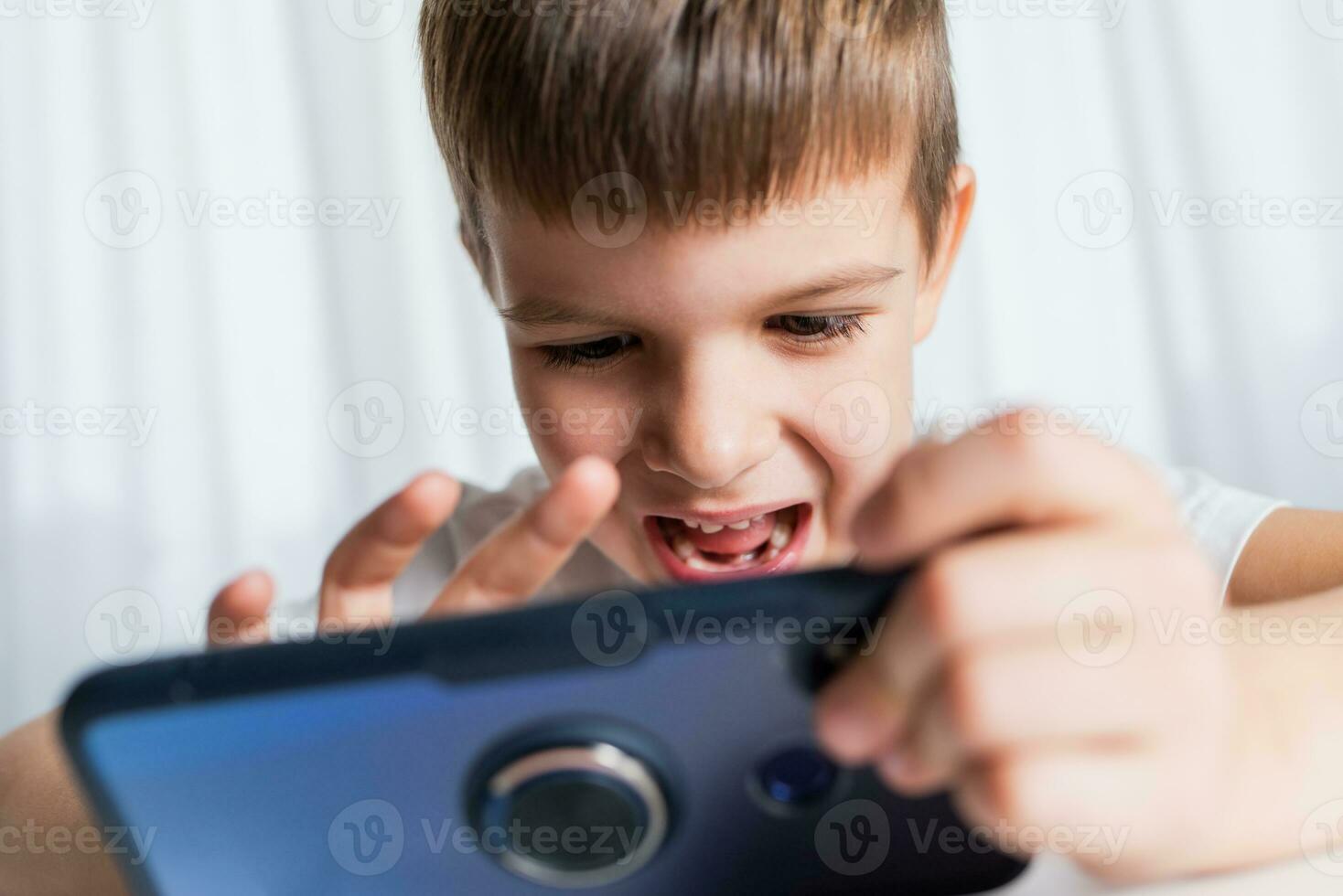 une de bonne humeur enfant dans une blanc T-shirt pièces Jeux sur le téléphone à maison. content garçon regards à le sien téléphone intelligent. photo