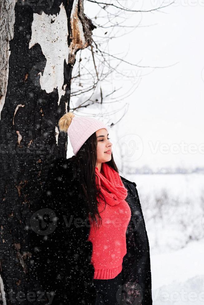 Portrait d'une jeune femme souriante debout près de l'arbre dans les chutes de neige photo