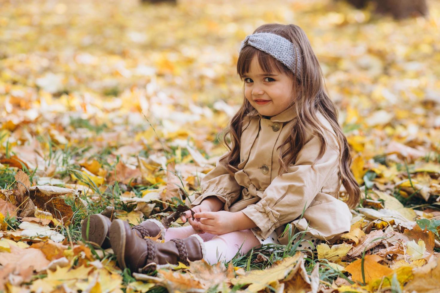 petite fille dans un manteau beige assise parmi les feuilles dans le parc en automne. photo