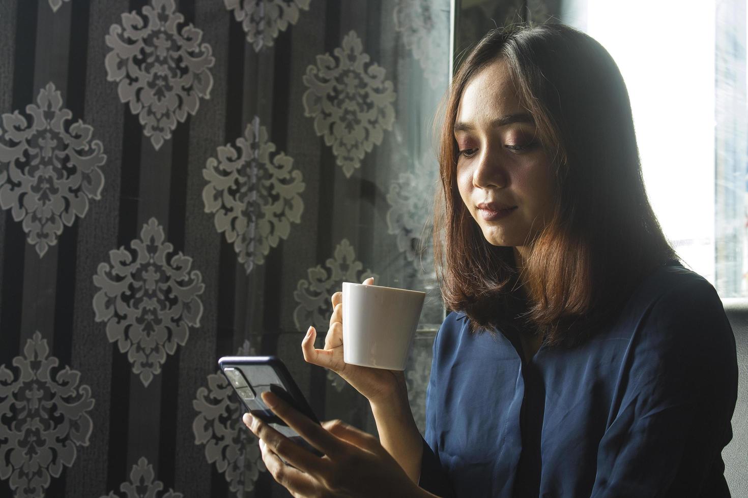 femme asiatique détendue buvant du café tout en utilisant un téléphone portable pour le travail photo