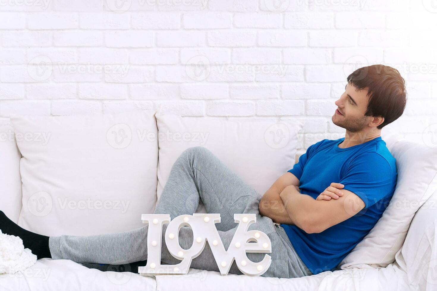 Beau Jeune homme bras franchi séance sur blanc canapé avec l'amour alphabet à la recherche à quelque chose. l'amour et la Saint-Valentin journée concept. photo
