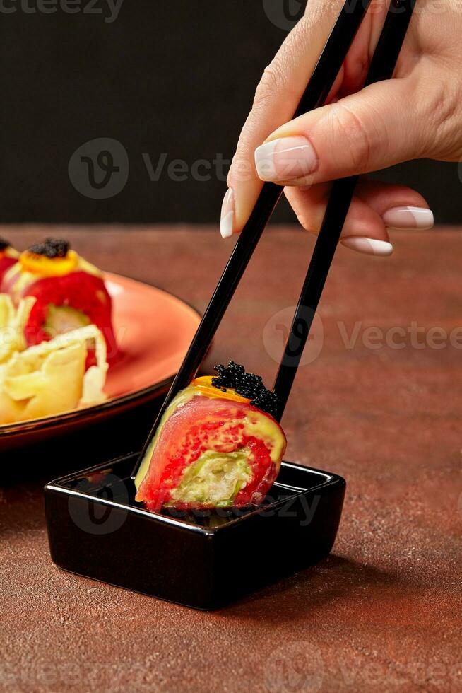 femelle main avec baguettes plongement rouge riz Sushi rouleau avec thon, noir caviar et kumquat dans soja sauce photo