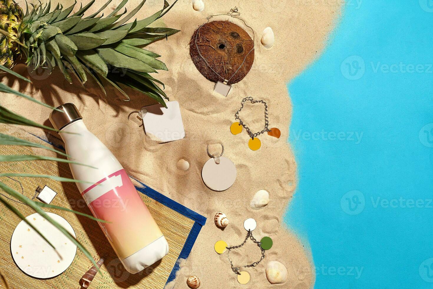 bijoux, cosmétique bouteille avec non étiqueter, coquilles, noix de coco, ananas, paume brindille et porte-clés sur le sable et bleu Contexte. proche en haut, copie espace photo